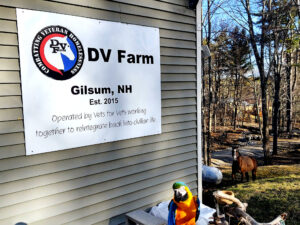 Outside of DV Farm office where there is a sign that says DV Farm, Gilsum New Hampshire and Operated by vets for vets working together to reintegrate back into civilian life. Ashe the horse is in the background and Gabby Sue the Macaw sits almost center and bottom of the image.